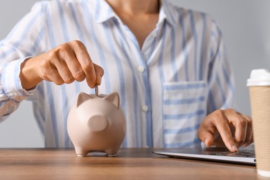 Photo of Woman putting coin into piggy bank while using laptop at wooden table, closeup