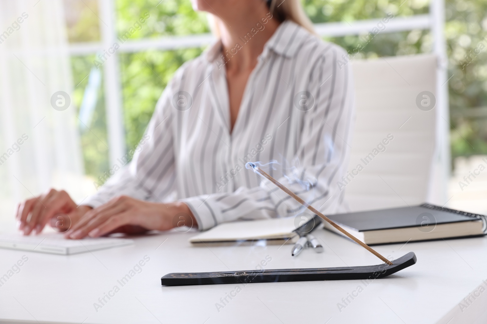 Photo of Woman working with computer at table while incense stick smoldering in holder indoors, selective focus