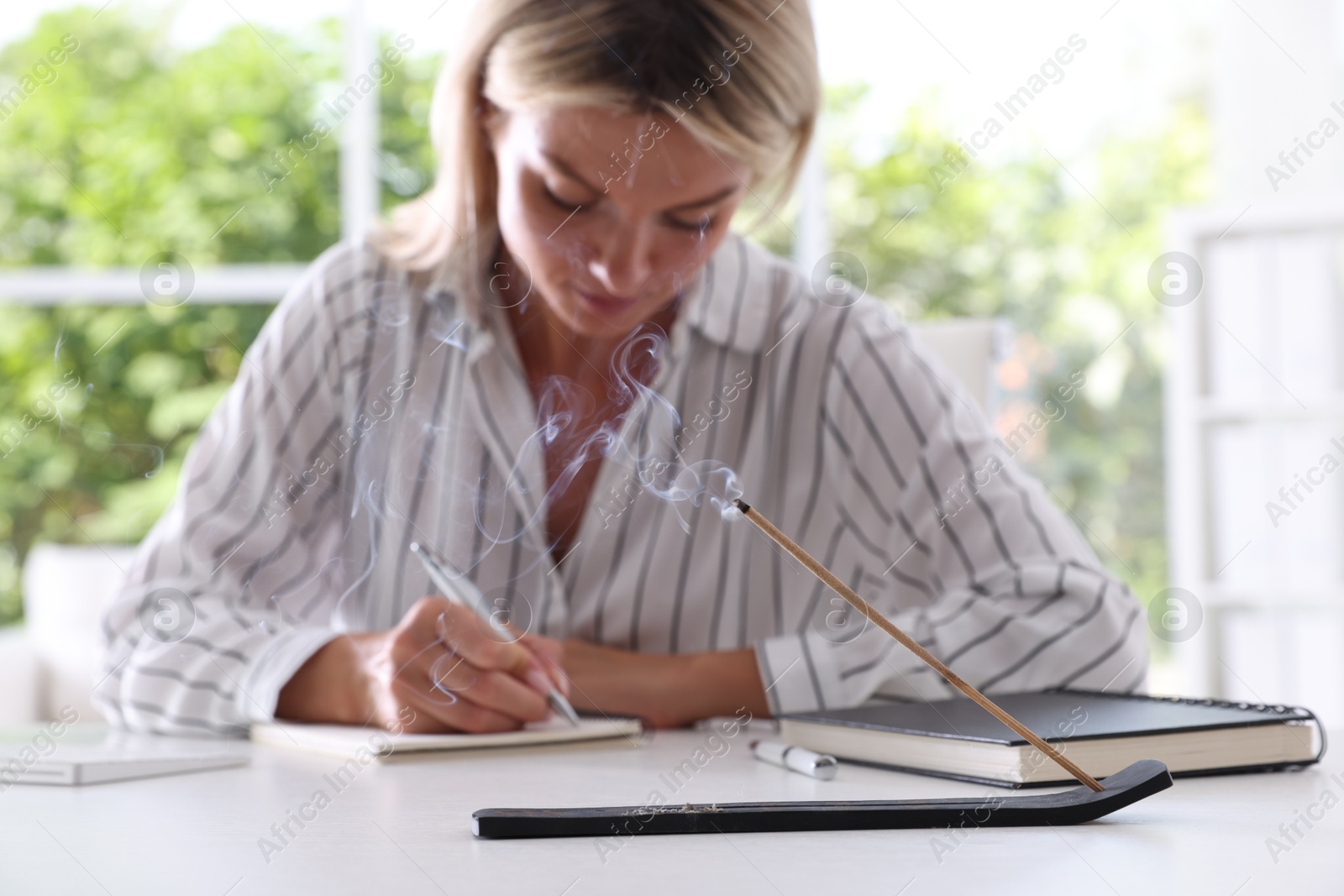 Photo of Woman working at table while incense stick smoldering in holder indoors, selective focus