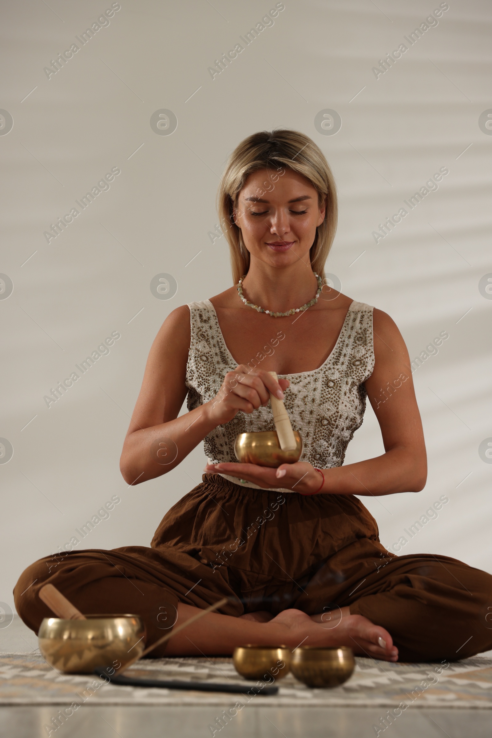 Photo of Beautiful young woman with smoldering incense stick and tibetan singing bowls on floor indoors