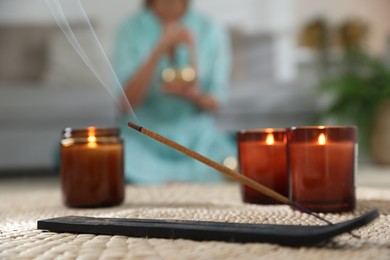 Incense stick smoldering in holder and burning candles on wicker mat indoors, selective focus