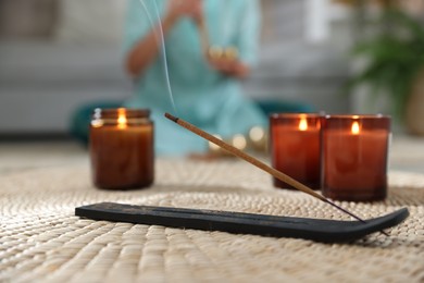 Incense stick smoldering in holder and burning candles on wicker mat indoors, selective focus