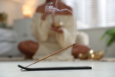 Incense stick smoldering in holder on floor indoors, selective focus