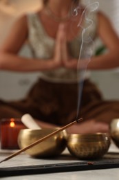 Young woman practicing yoga indoors, focus on smoldering incense stick and tibetan singing bowls