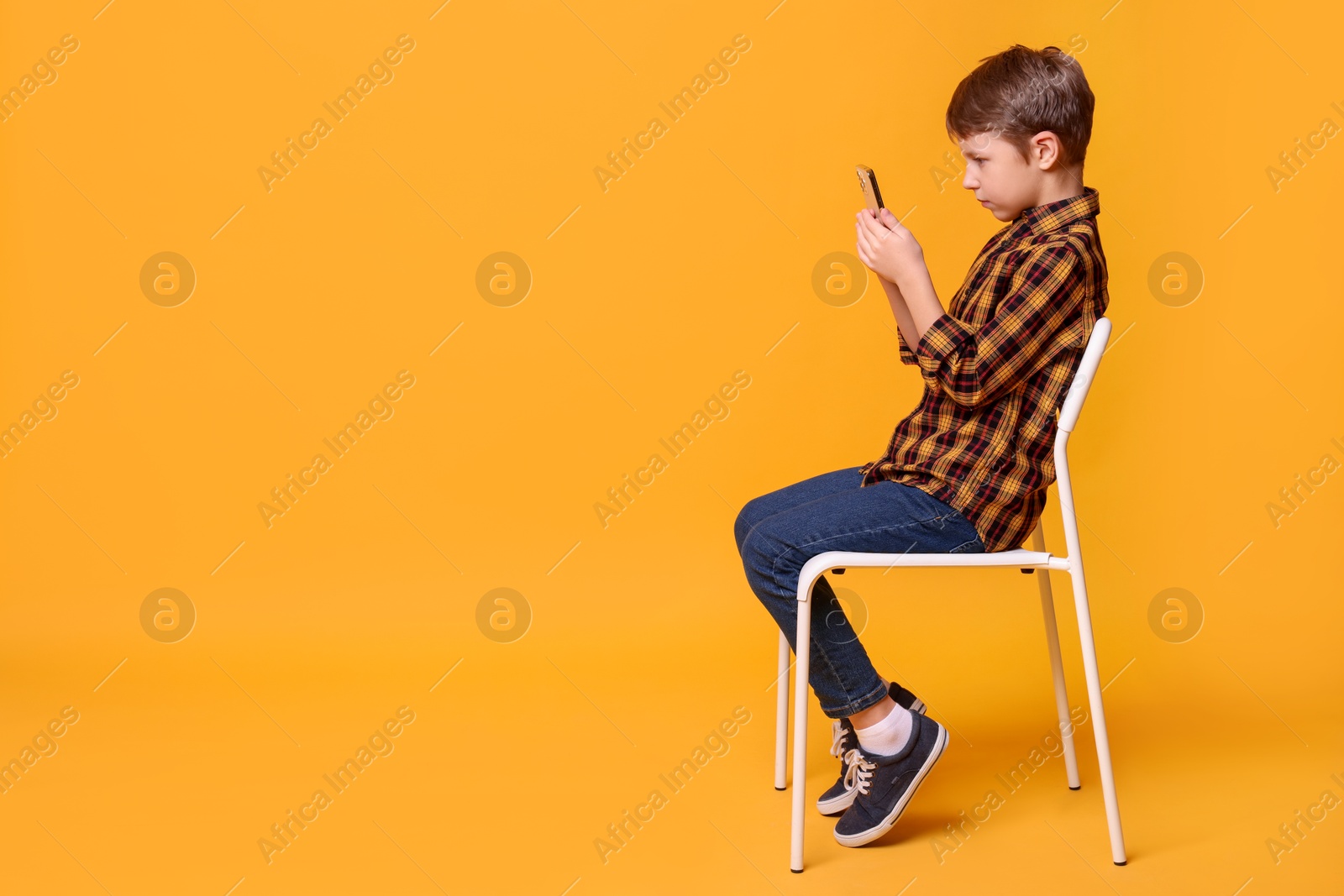 Photo of Boy with correct posture and smartphone sitting on chair against yellow background