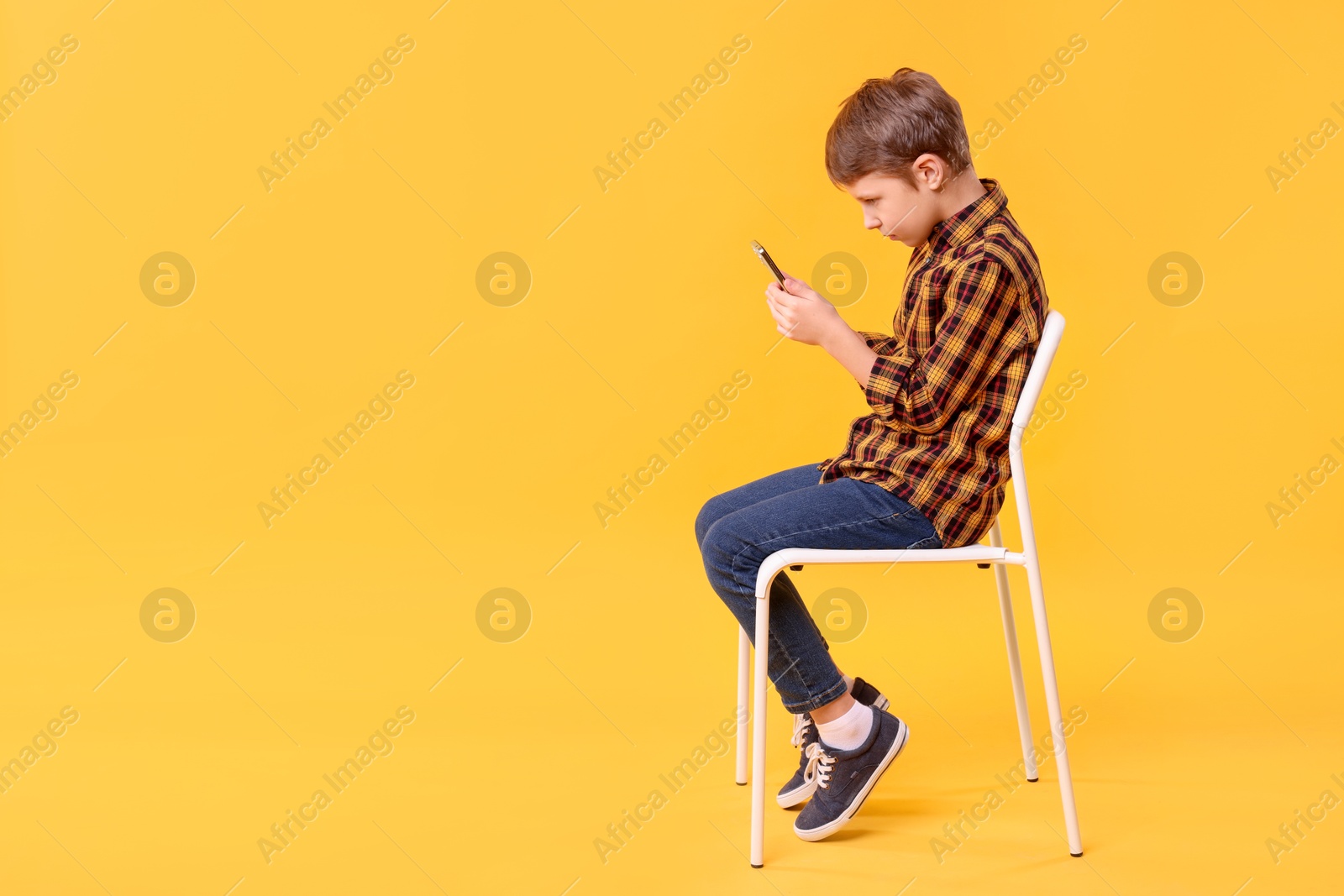 Photo of Boy with incorrect posture and smartphone sitting on chair against yellow background