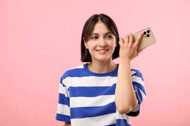 Beautiful woman with smartphone listening to voice message on pink background