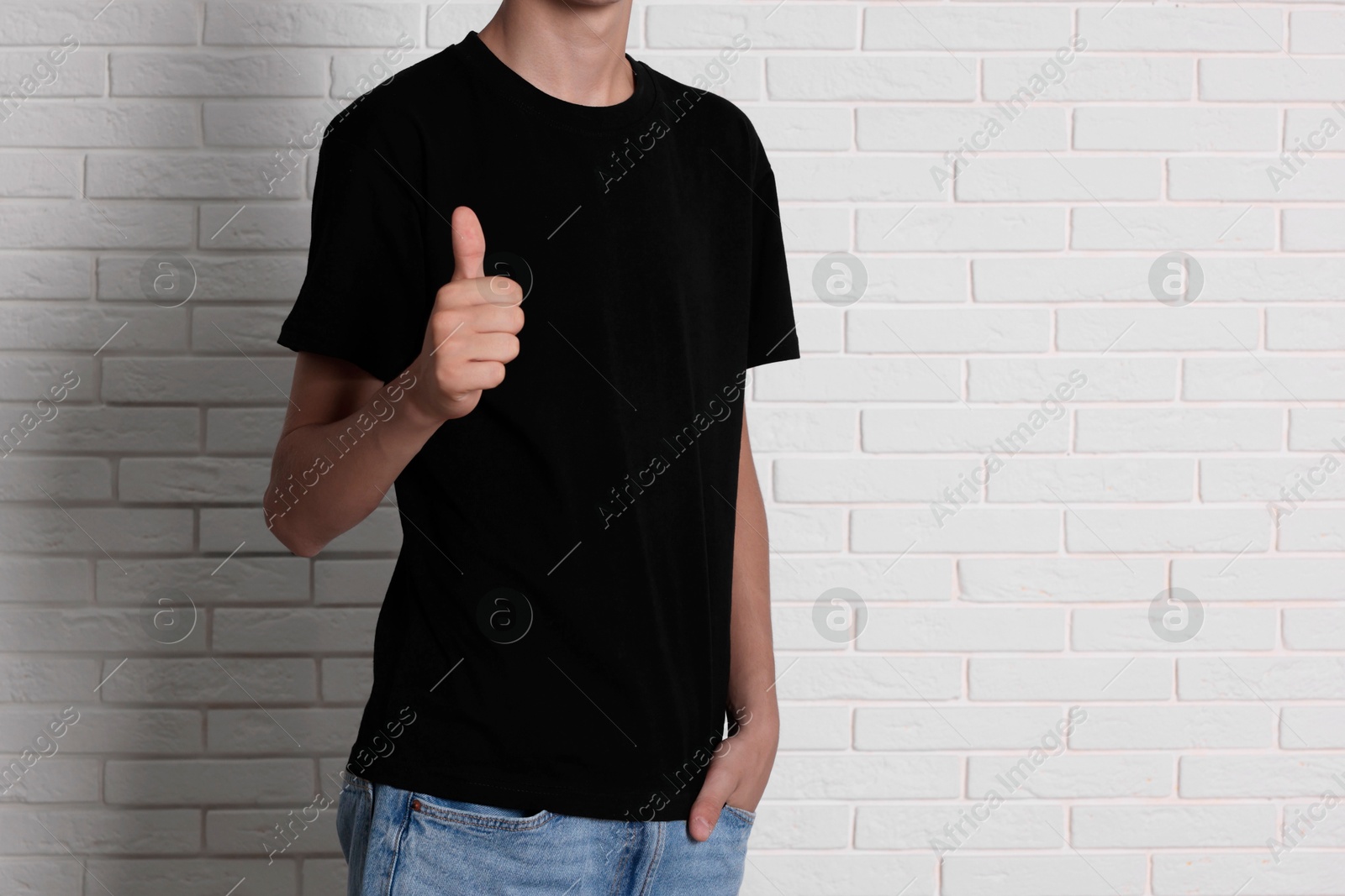 Photo of Teenage boy wearing black t-shirt and showing thumbs up near white brick wall, closeup. Space for text