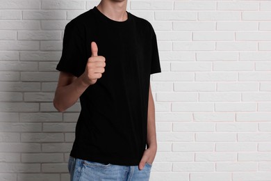 Photo of Teenage boy wearing black t-shirt and showing thumbs up near white brick wall, closeup. Space for text