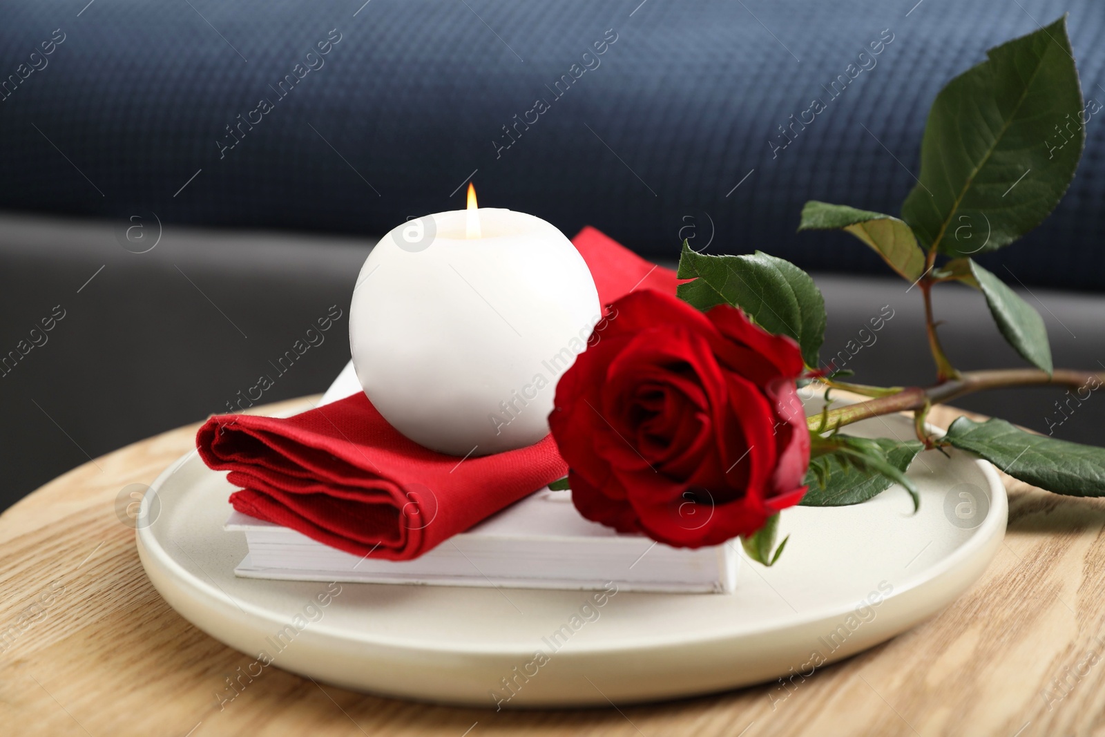 Photo of Burning candle, napkin, red rose and book on wooden table indoors, closeup