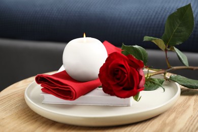 Photo of Burning candle, napkin, red rose and book on wooden table indoors, closeup