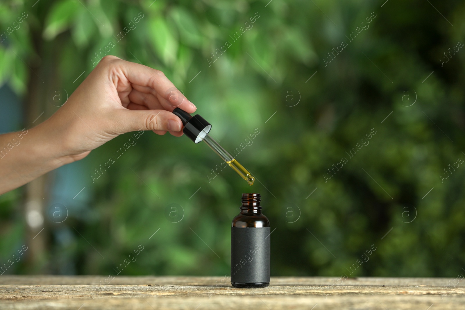 Photo of Woman dripping tincture from pipette into bottle at wooden table against blurred green background, closeup