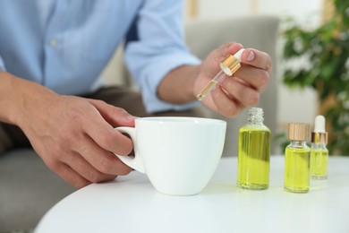 Photo of Young man dripping CBD tincture into drink at white table, closeup