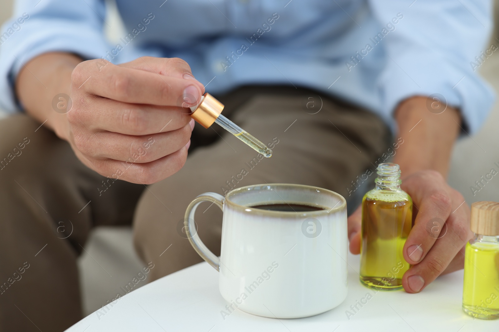 Photo of Young man dripping CBD tincture into drink at white table, closeup