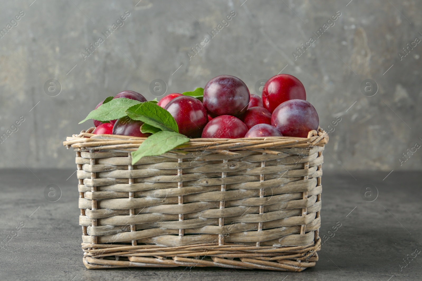 Photo of Tasty ripe plums and leaves in wicker basket on grey textured table