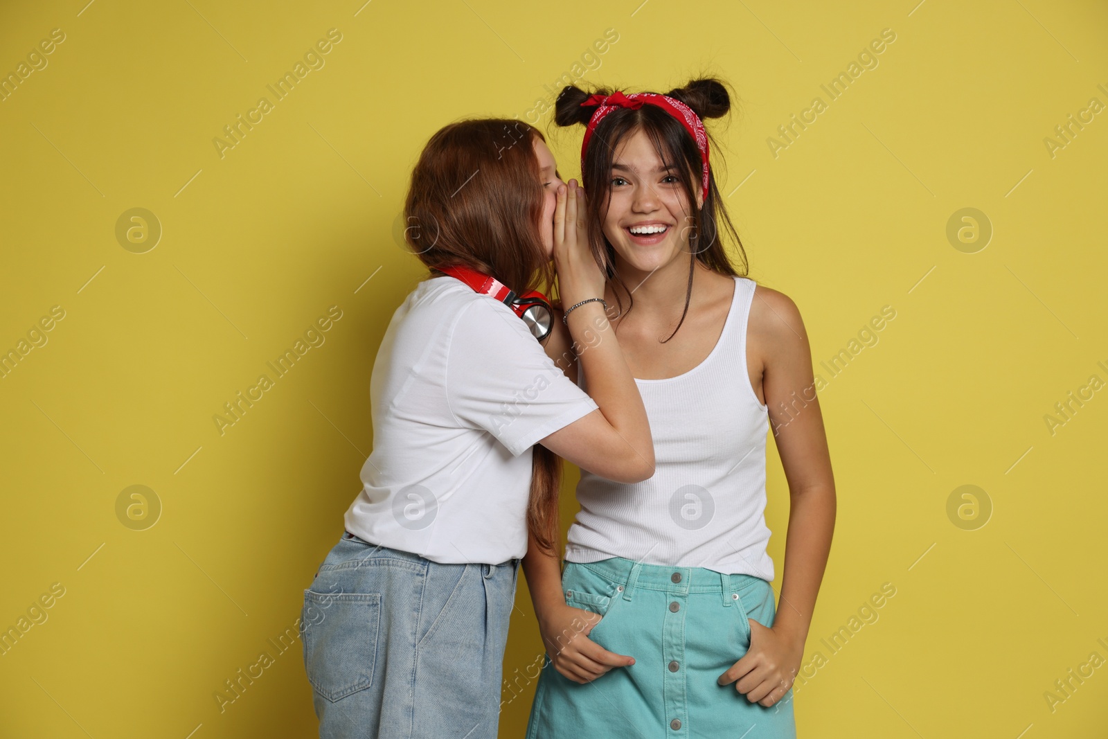 Photo of Teenage girl whispering secret to her happy friend on yellow background