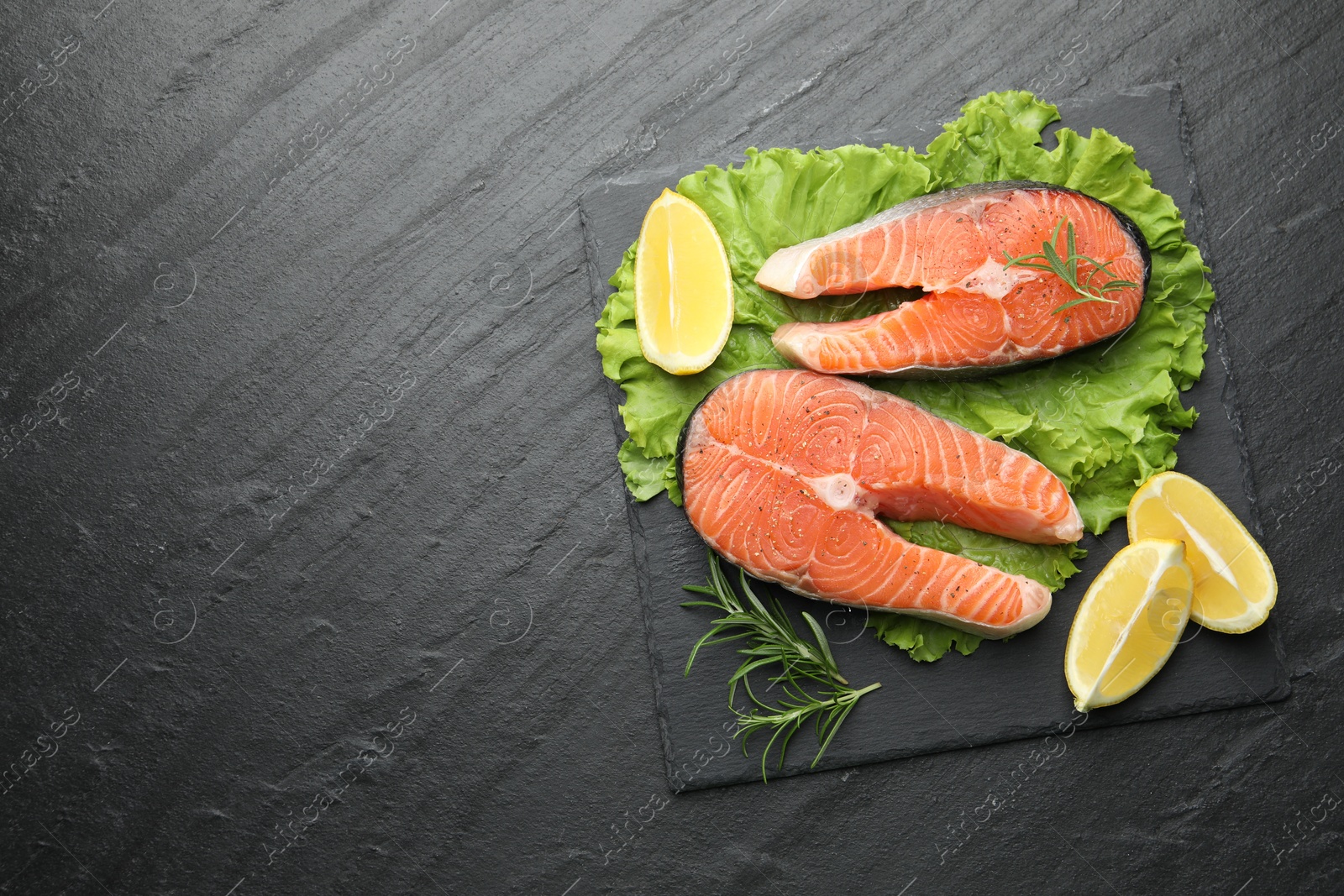 Photo of Fresh raw salmon steaks with spices, lemon, lettuce and rosemary on dark textured table, top view. Space for text