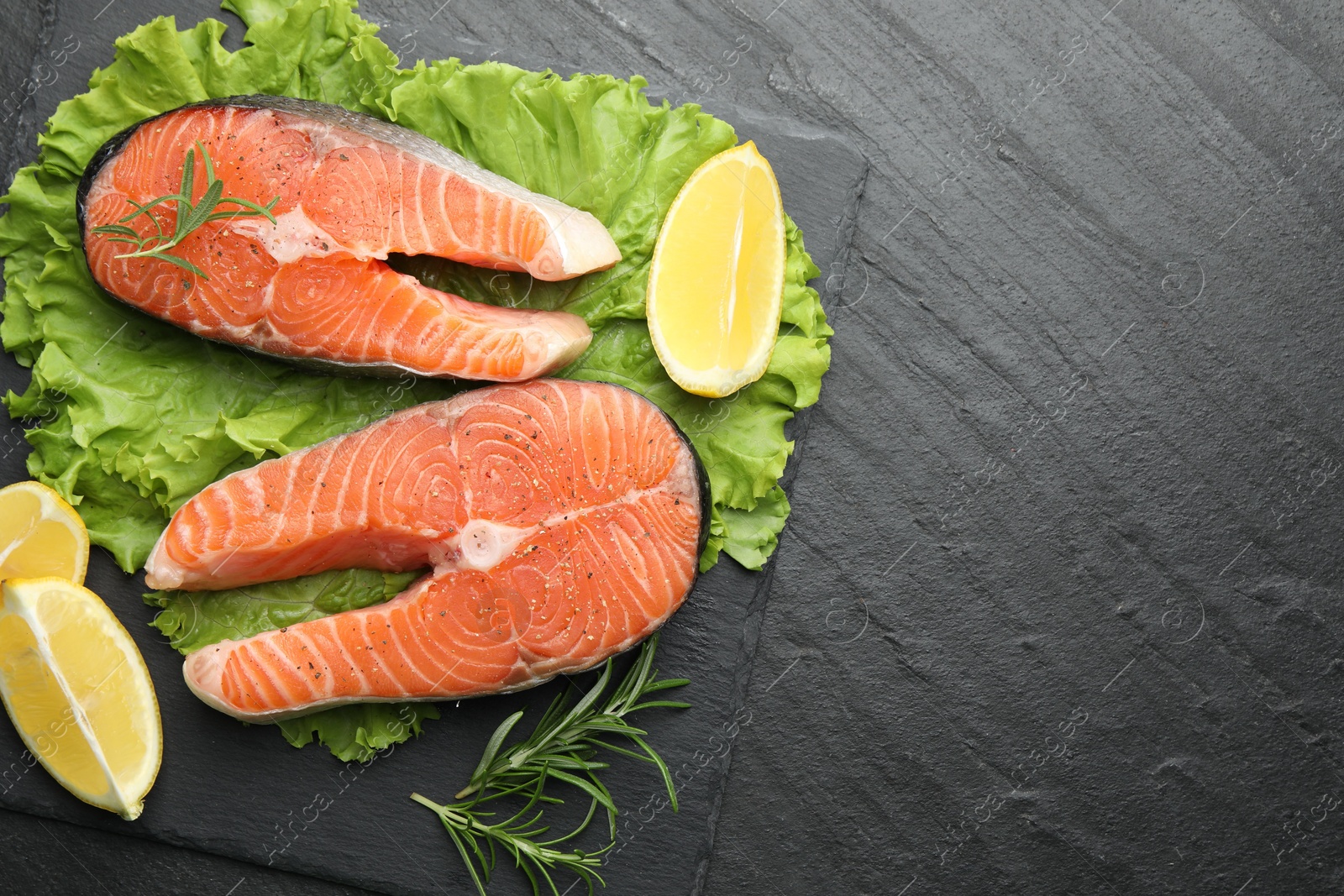 Photo of Fresh raw salmon steaks with spices, lemon, lettuce and rosemary on dark textured table, top view. Space for text