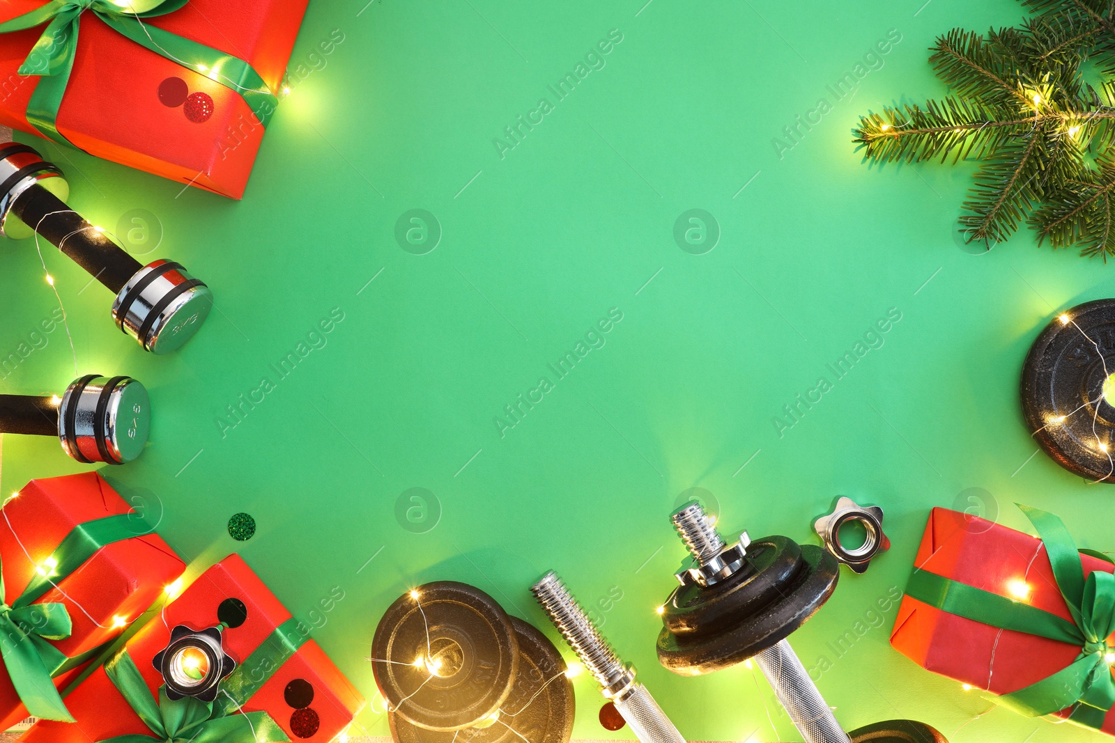 Photo of Dumbbells, fir tree branches, gift boxes and Christmas lights on green background, flat lay. Space for text