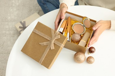 Photo of Woman holding Christmas gift box with makeup brushes and cosmetics at white table, closeup