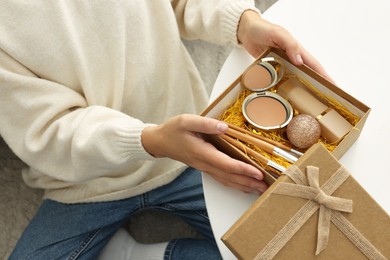 Photo of Woman holding Christmas gift box with makeup brushes and cosmetics at white table, closeup