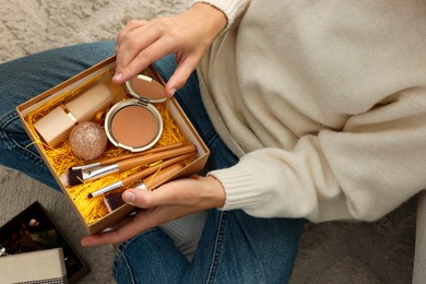 Photo of Woman holding Christmas gift box with makeup brushes and cosmetics at home, above view