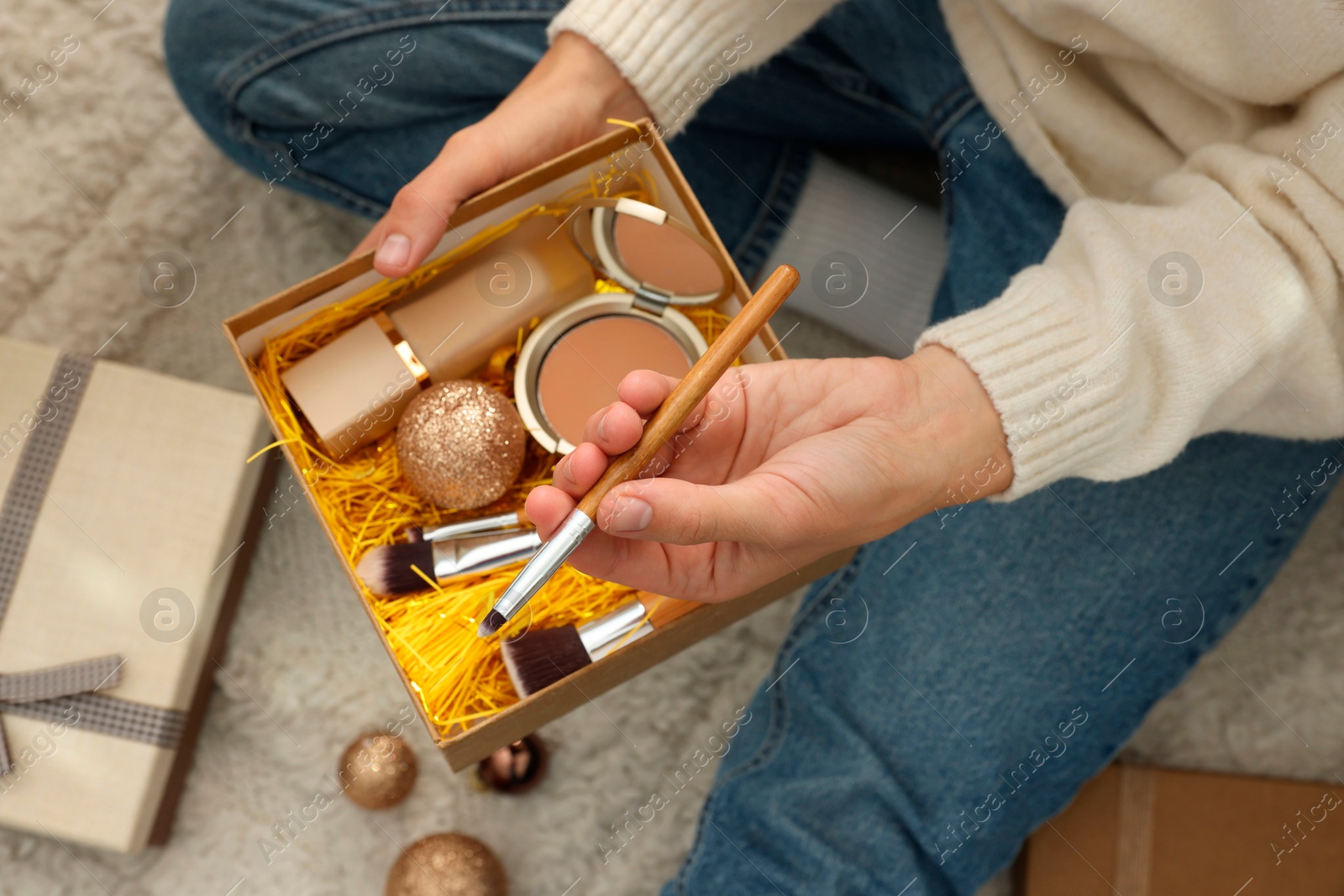 Photo of Woman holding Christmas gift boxes with makeup brushes and cosmetics at home, above view