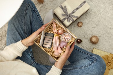 Photo of Woman holding Christmas gift boxes with makeup brushes and cosmetics at home, above view