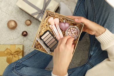 Photo of Woman holding Christmas gift boxes with makeup brushes and cosmetics at home, above view