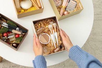 Photo of Woman holding Christmas gift boxes with makeup brushes and cosmetics at home, above view