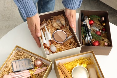 Photo of Woman holding Christmas gift boxes with makeup brushes and cosmetics at white table, closeup