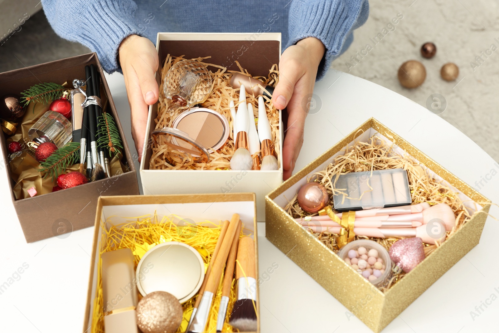 Photo of Woman holding Christmas gift boxes with makeup brushes and cosmetics at white table, closeup