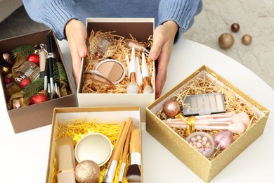 Photo of Woman holding Christmas gift boxes with makeup brushes and cosmetics at white table, closeup