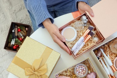 Photo of Woman holding Christmas gift boxes with makeup brushes and cosmetics at white table, above view