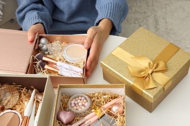 Photo of Woman holding Christmas gift boxes with makeup brushes and cosmetics at white table, closeup