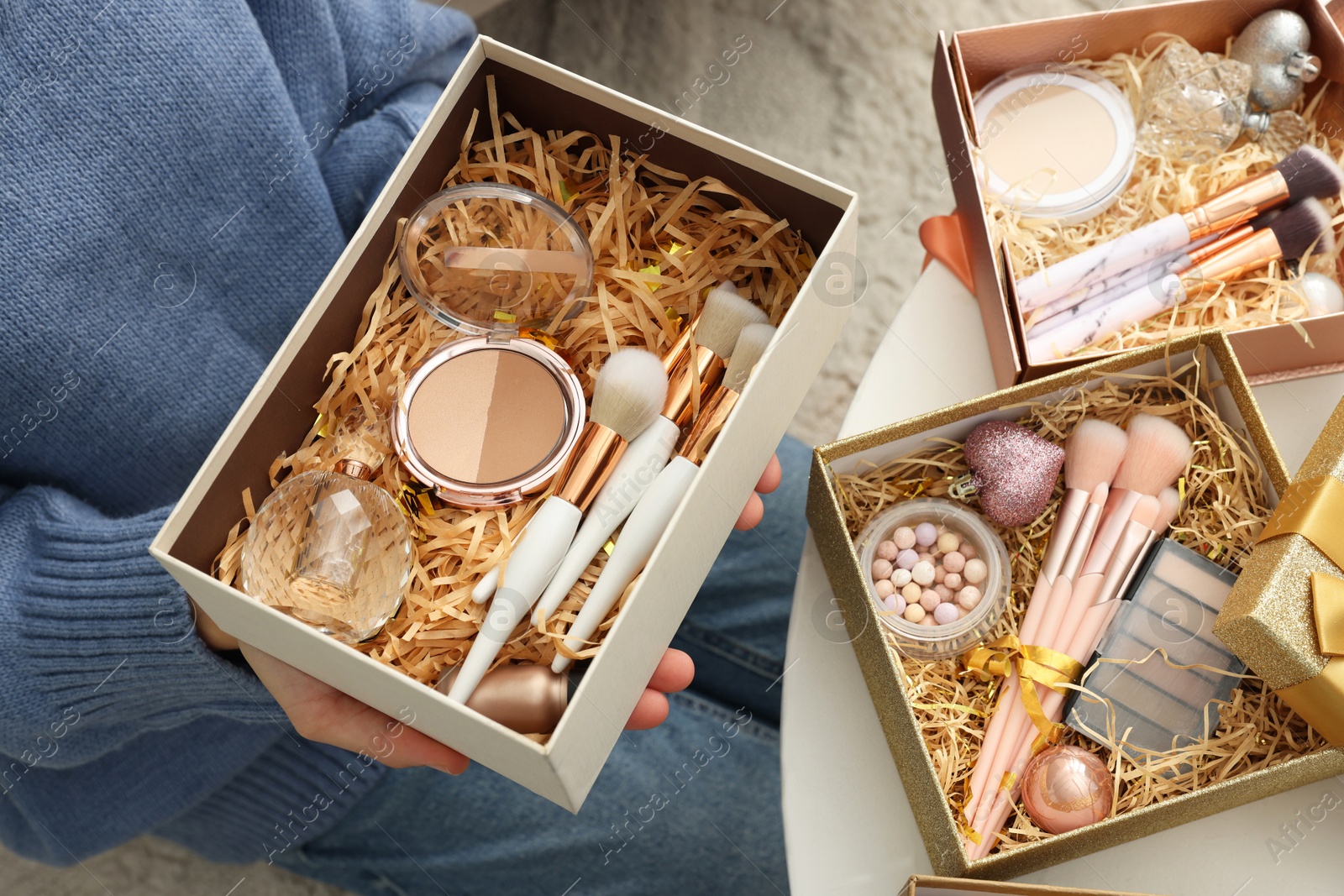 Photo of Woman holding Christmas gift boxes with makeup brushes and cosmetics at white table, closeup