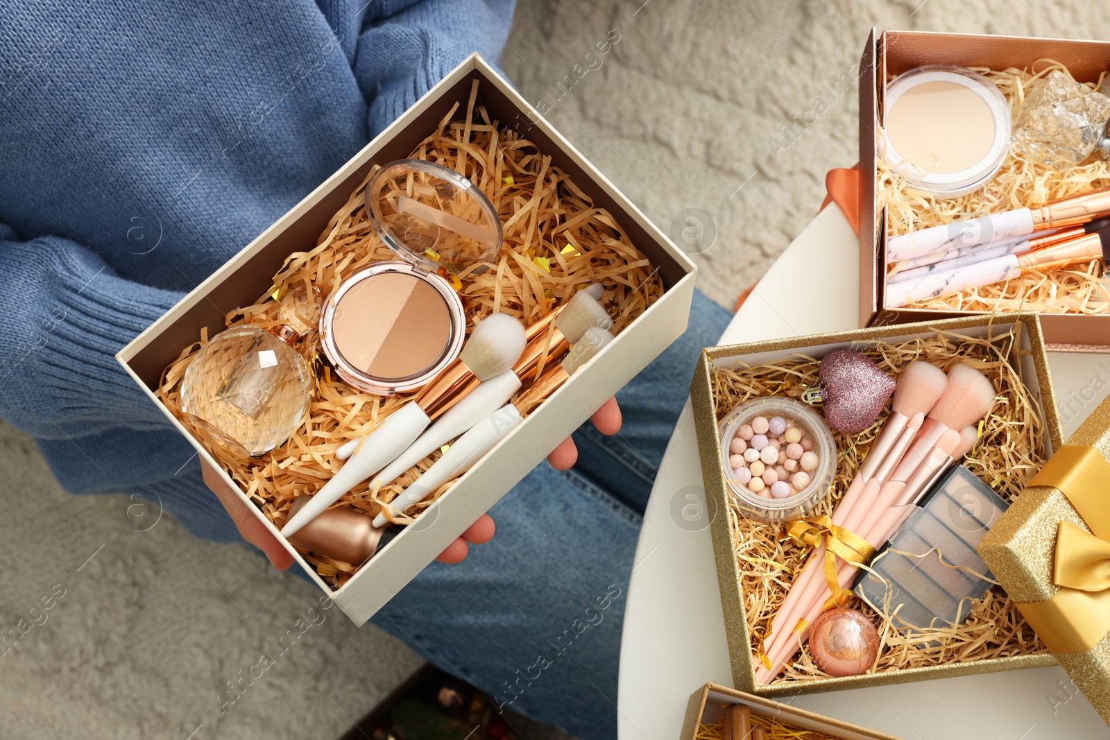 Photo of Woman holding Christmas gift boxes with makeup brushes and cosmetics at white table, above view