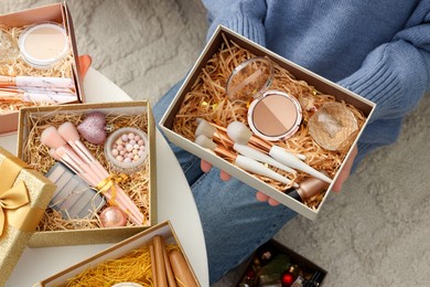 Photo of Woman holding Christmas gift boxes with makeup brushes and cosmetics at white table, above view