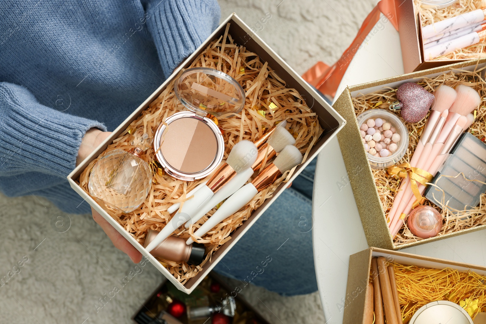 Photo of Woman holding Christmas gift boxes with makeup brushes and cosmetics at white table, above view