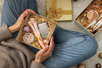 Photo of Woman holding Christmas gift boxes with makeup brushes and cosmetics at home, above view