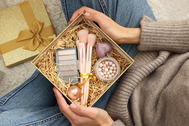 Photo of Woman holding Christmas gift boxes with makeup brushes and cosmetics at home, above view