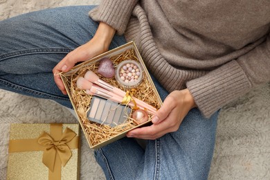 Photo of Woman holding Christmas gift box with makeup brushes and cosmetics at home, above view