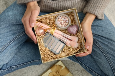 Photo of Woman holding Christmas gift box with makeup brushes and cosmetics at home, above view