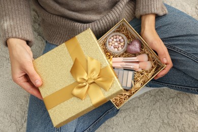 Photo of Woman holding Christmas gift box with makeup brushes and cosmetics at home, above view