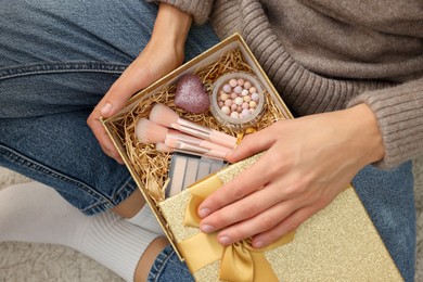 Photo of Woman holding Christmas gift box with makeup brushes and cosmetics at home, above view