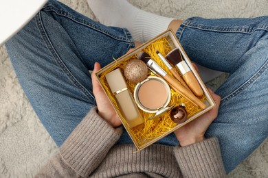 Photo of Woman holding Christmas gift box with makeup brushes and cosmetics at home, above view