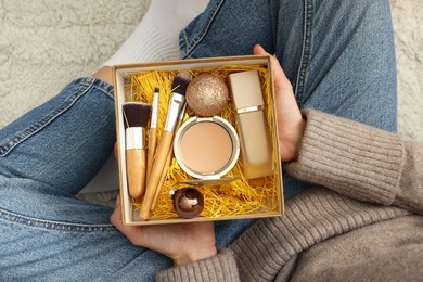 Photo of Woman holding Christmas gift box with makeup brushes and cosmetics at home, above view