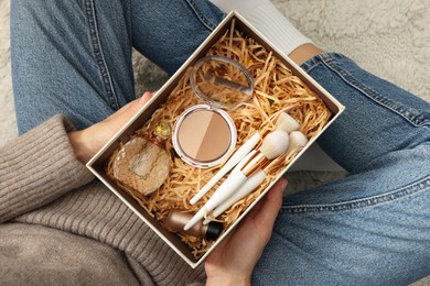 Photo of Woman holding Christmas gift box with makeup brushes and cosmetics at home, above view