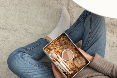 Photo of Woman holding Christmas gift box with makeup brushes and cosmetics at home, above view