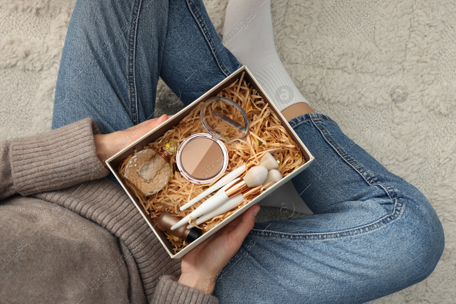 Photo of Woman holding Christmas gift box with makeup brushes and cosmetics at home, above view